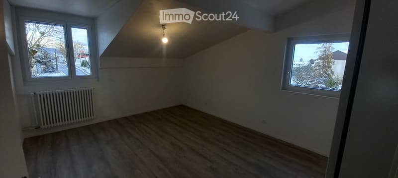 Empty room with wooden flooring, white walls, and large windows offering a view of the snowy landscape outside. A radiator is visible on one wall, and the ceiling has a sloped design with a single light fixture.