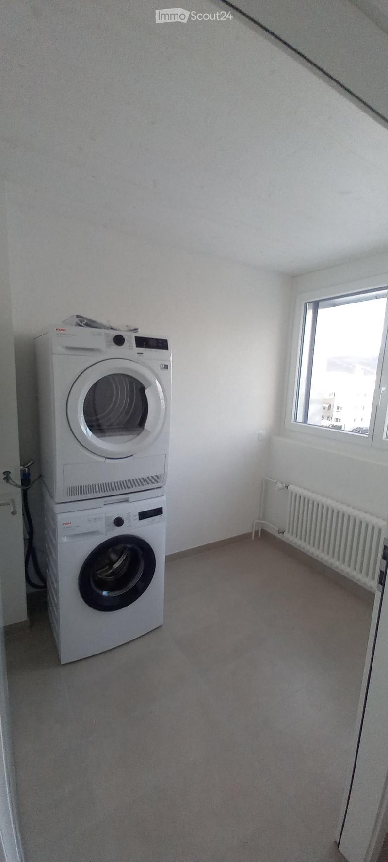 Small room with a stacked washing machine and dryer unit against one wall, a window on the opposite wall, and a radiator below it.