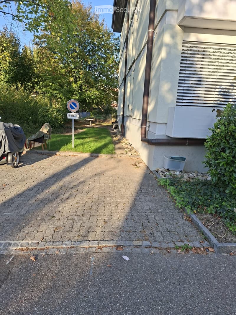 A paved driveway leads to a house with a covered car parked on the left. The house has white walls, a visible air conditioning unit, and a small garden area with plants and a covered chair. A "PRIVAT" sign is visible near the driveway.