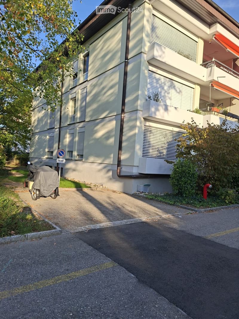 Two-story house with light-colored facade, multiple windows, and a covered balcony area on the upper floor. A scooter is parked near the entrance, and the driveway is paved.
