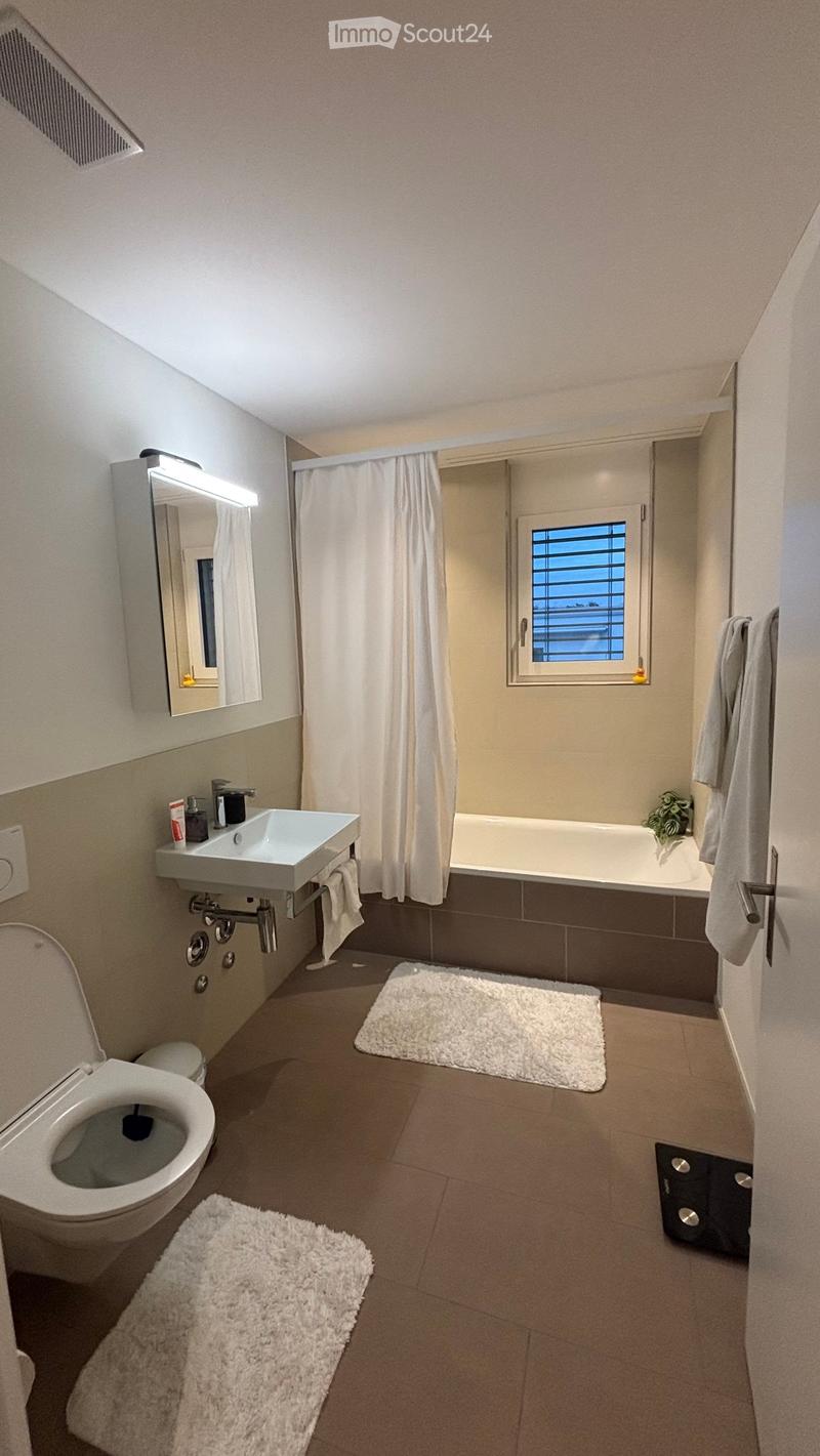 Modern bathroom with a toilet, sink, bathtub, mirror, and towels. Features a window with a view, a shower curtain, and two white rugs on the floor.