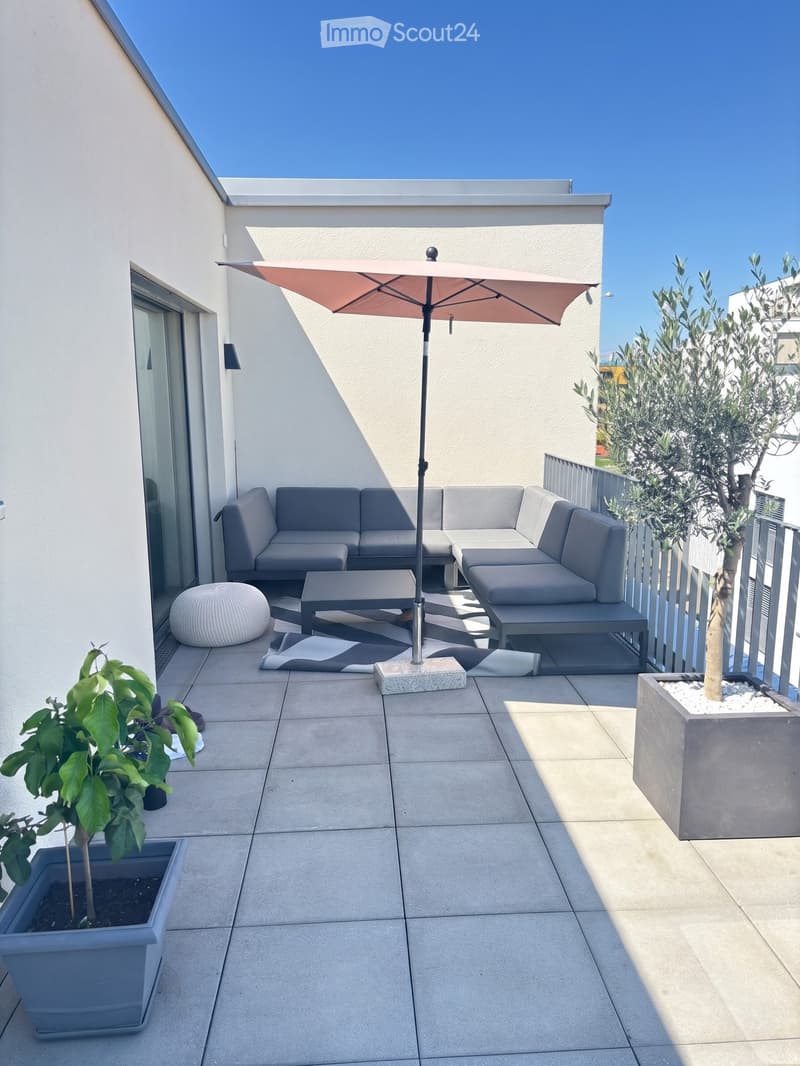 Modern balcony with grey sectional sofa, coffee table, umbrella, and potted plants.