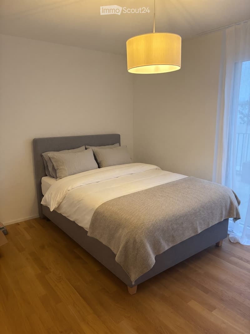 A modern bedroom with a neatly made double bed, light-colored bedding, and a grey headboard. The room features wooden flooring and a pendant light hanging from the ceiling.