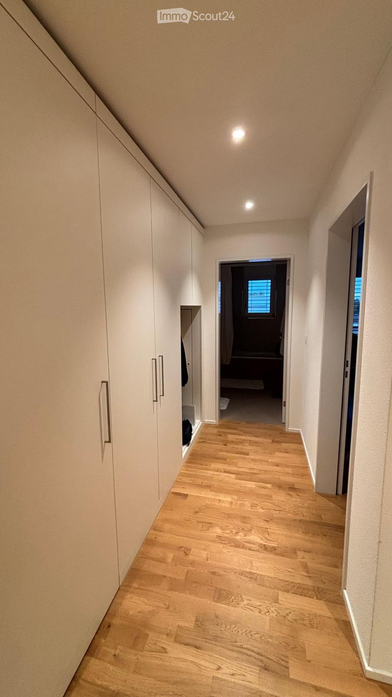 A modern hallway with light wooden flooring, white walls, built-in white cabinets with silver handles, and recessed ceiling lighting. A doorway leads to another room with a visible window and a glimpse of furniture.
