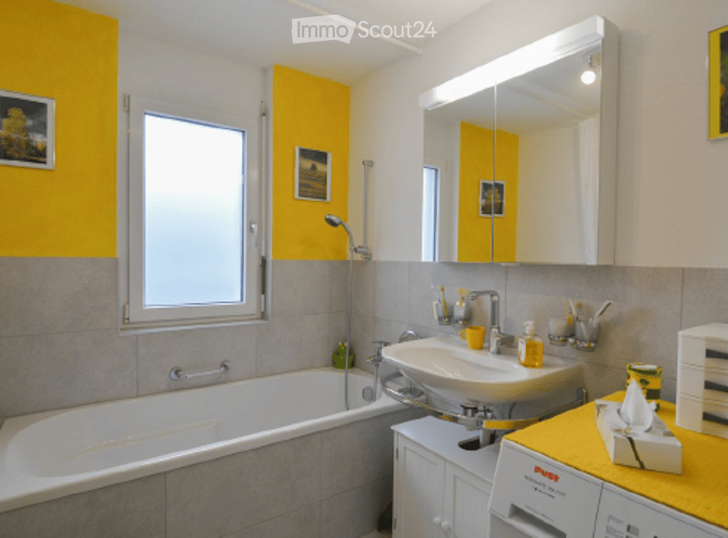 Bright bathroom with yellow accent wall, white tiles, bathtub, sink, mirror, and various toiletries.