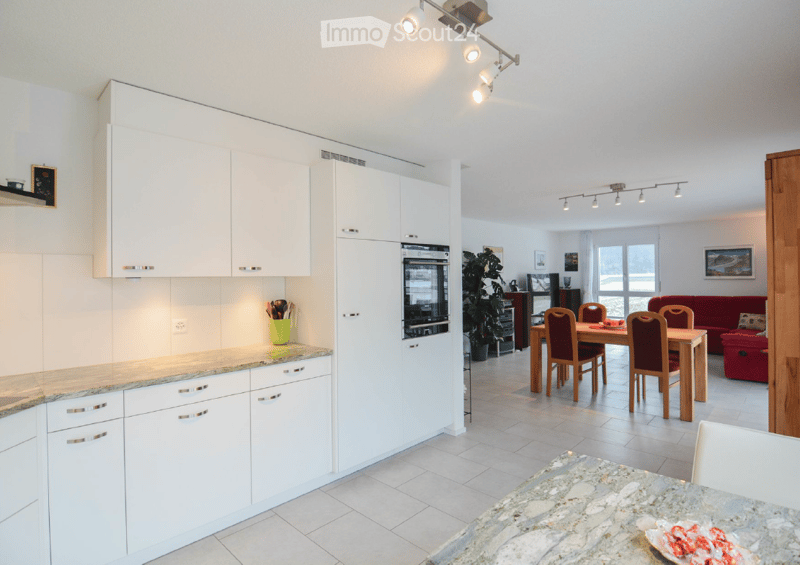 Open-plan kitchen and living area with white cabinetry, granite countertops, a dining table with chairs, a red sofa, and a TV in the background.