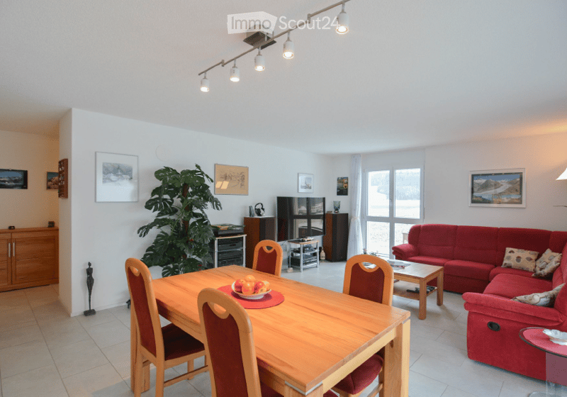 Bright living and dining area with a red sofa, wooden dining table, chairs, coffee table, TV stand, and decorative plant. The space features large windows and modern lighting.