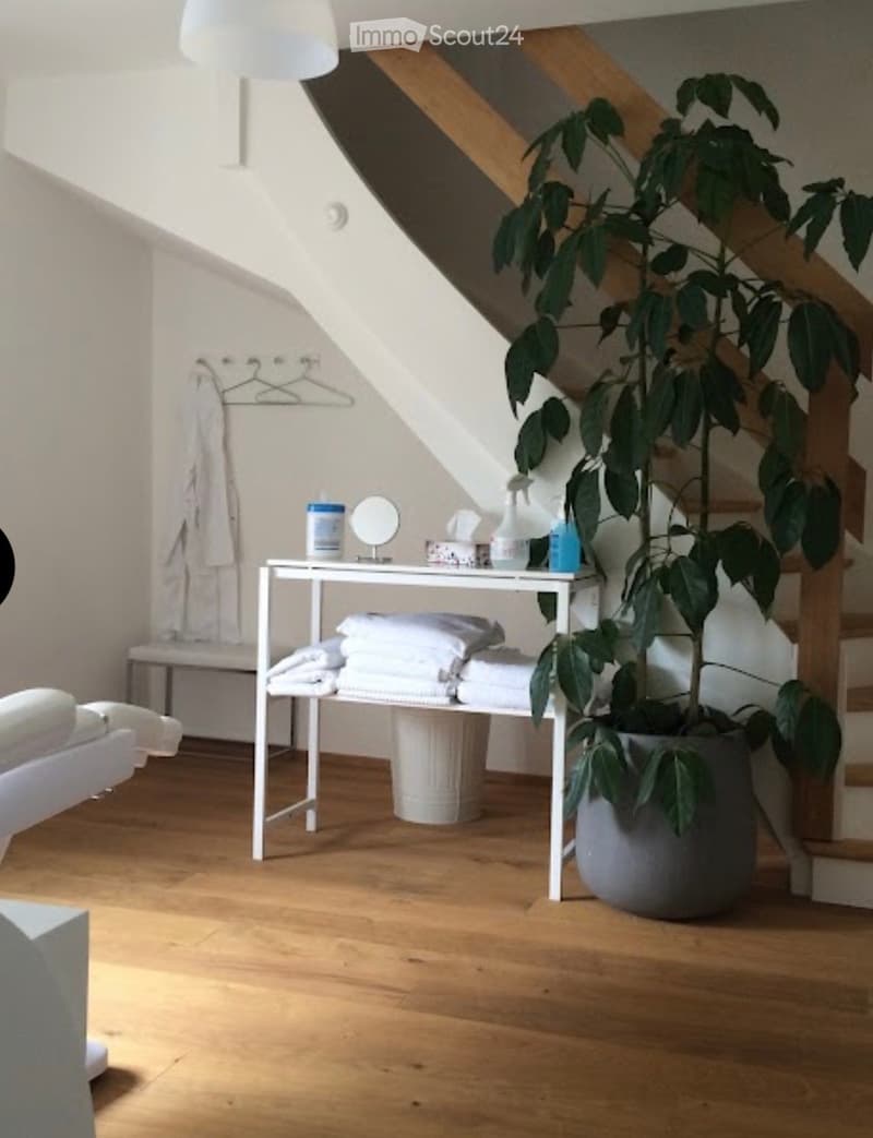 A cozy, well-lit room with wooden flooring, a large potted plant, a small table with towels and a mirror, and a staircase leading upwards.