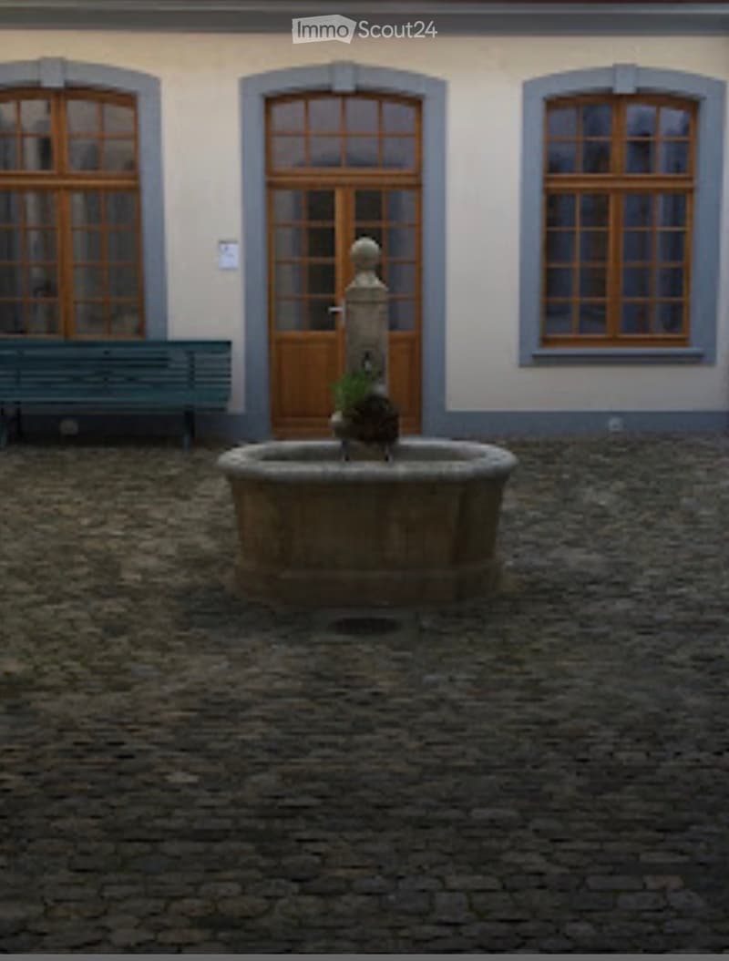 Courtyard with a stone fountain, wooden bench, and building with large windows and doors.