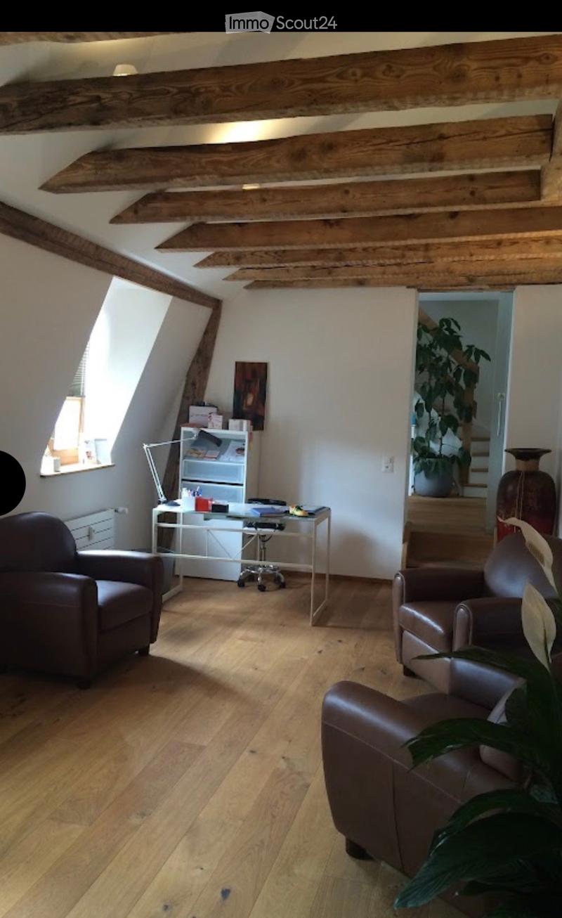 Cozy attic living space with wooden beams, hardwood floor, leather sofa, armchair, small table, bookshelf, and large window letting in natural light.