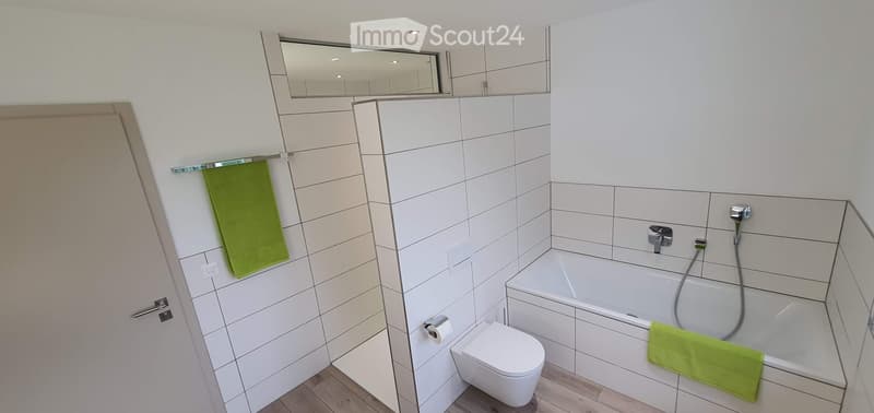 Modern bathroom with white tiles, bathtub, toilet, mirror, and green towels.