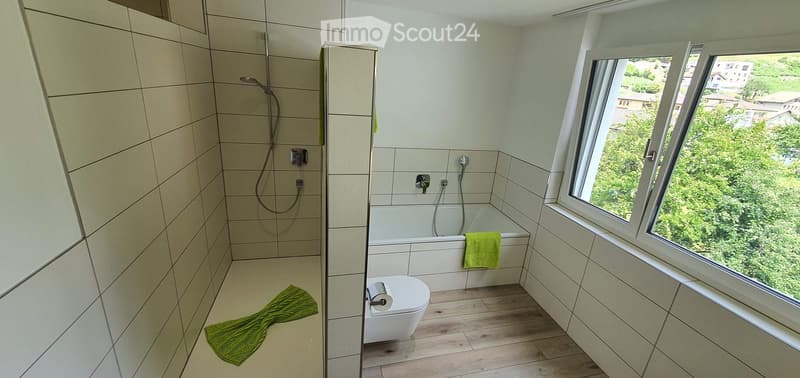 Modern bathroom with a bathtub, toilet, shower, and a large window offering a view of greenery outside. Green towels are placed on the floor and bathtub.