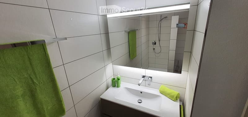 Modern bathroom with a white sink, chrome faucet, large mirror with built-in lighting, and green towels.