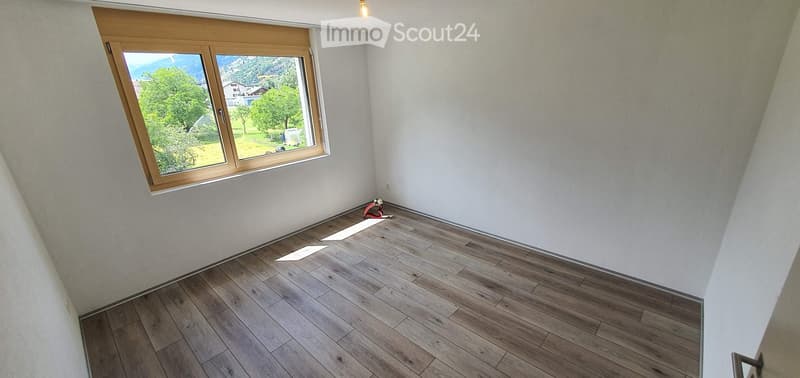 Empty room with wooden flooring and white walls, featuring a large window with wooden frame overlooking a green landscape.