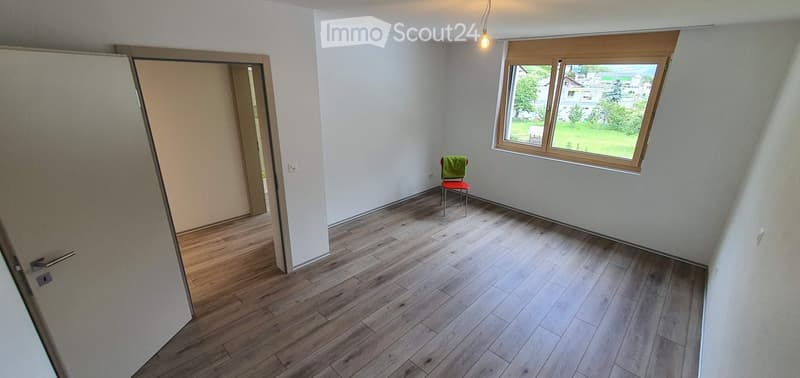 Empty room with wooden flooring, white walls, a window with a view, and a single chair with a green cushion.