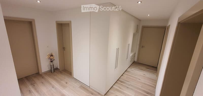 A clean, modern hallway with light wooden flooring, white walls, and built-in white closet units. A small vase with dried flowers sits on the floor near the left wall. Three doors are visible, two on the left and one on the right.