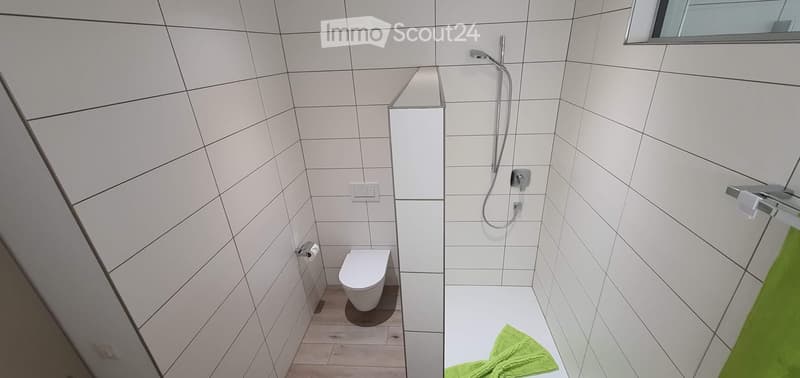 Modern bathroom with white tiled walls, a toilet, a shower area, and a green towel on the floor.