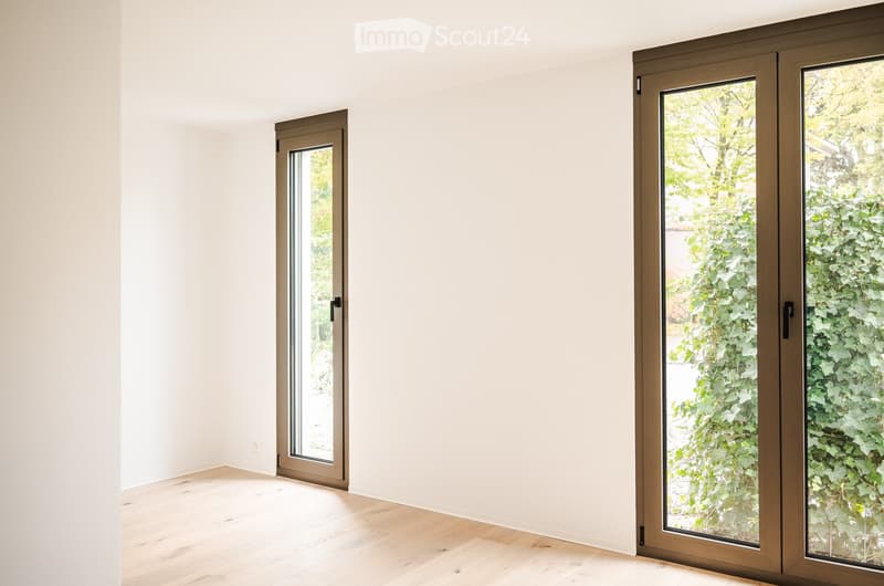 Empty room with light-colored wooden flooring and white walls, featuring two large sliding glass doors with brown frames that open to a green outdoor area.
