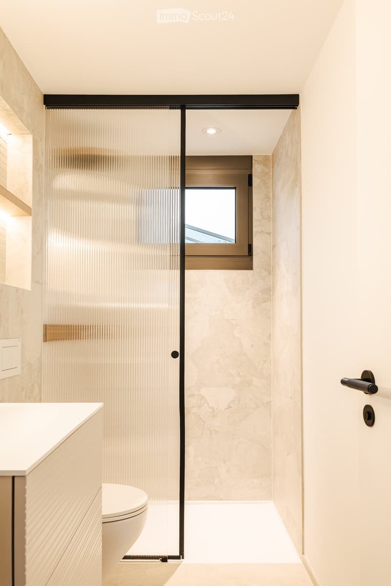 Modern bathroom with a white toilet, sink, and a glass shower enclosure with a frosted glass door. The walls are covered in marble tiles, and the floor is light-colored.