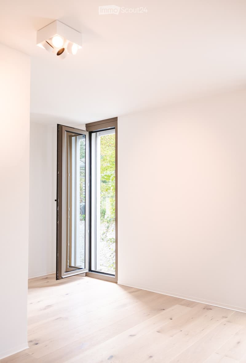 Empty room with light wooden flooring, white walls, and large sliding glass doors leading to a garden area.