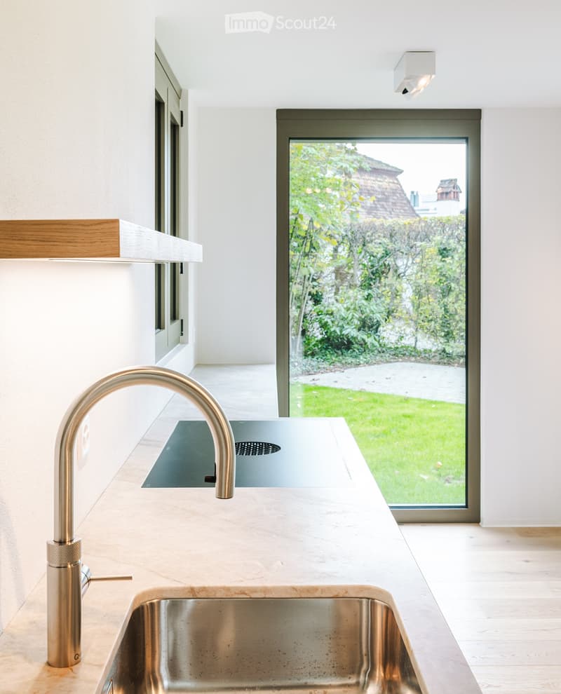 Modern kitchen with a sleek, minimalist design, featuring a stainless steel sink, a polished faucet, and a light-colored countertop. Large windows offer a view of a lush garden.