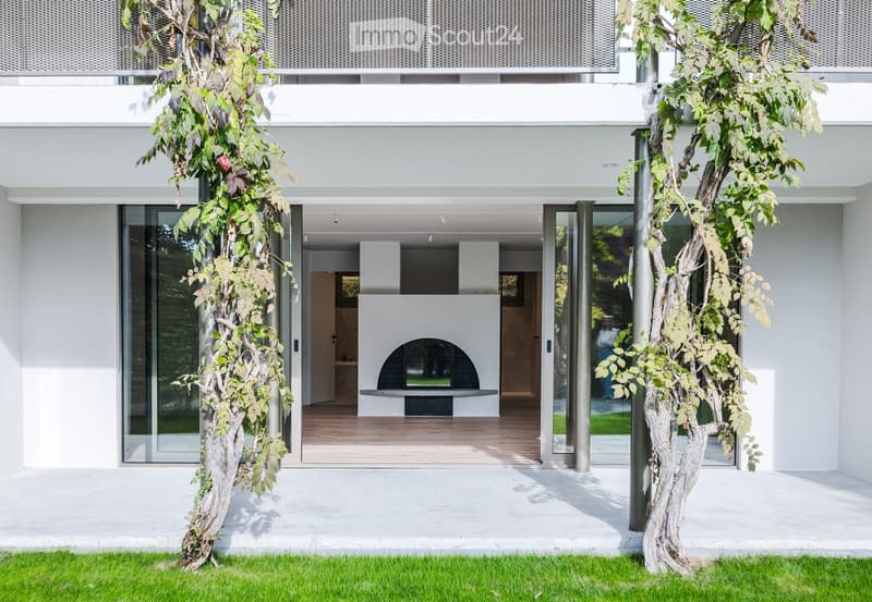 Modern white building with large glass doors leading to a patio area featuring a built-in fireplace and wooden flooring.
