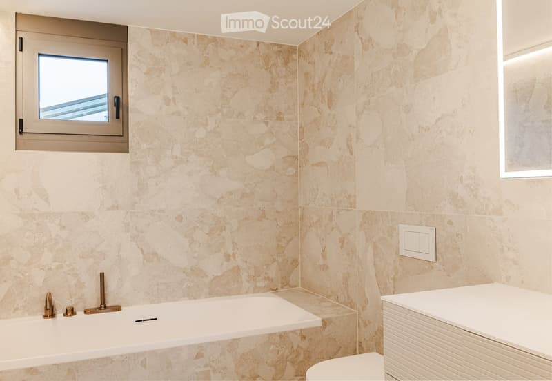 Modern bathroom with beige marble tiles, a white bathtub with brass fixtures, a white toilet, and a white cabinet.