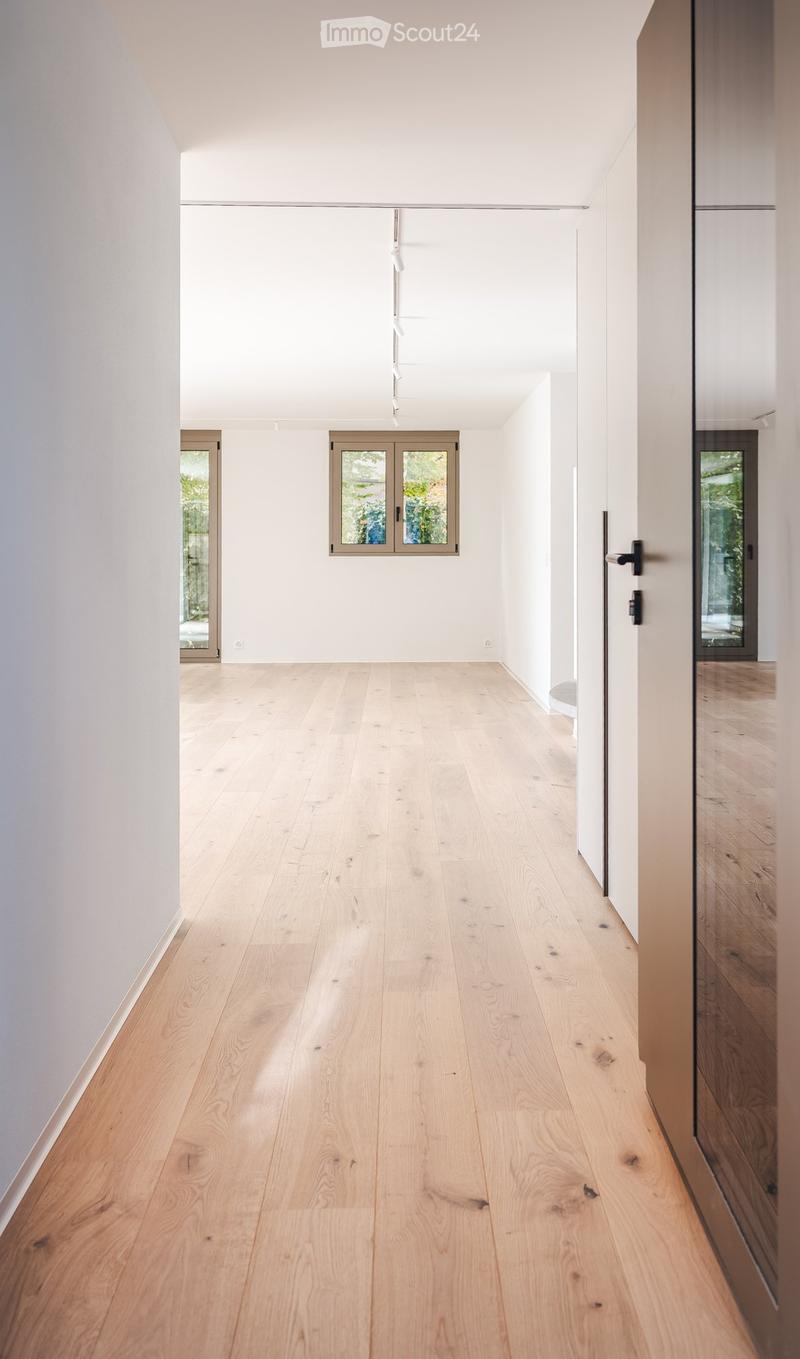 Bright, minimalist hallway with light wooden flooring, white walls, and large windows allowing natural light. Features modern glass doors and sleek black door handles.