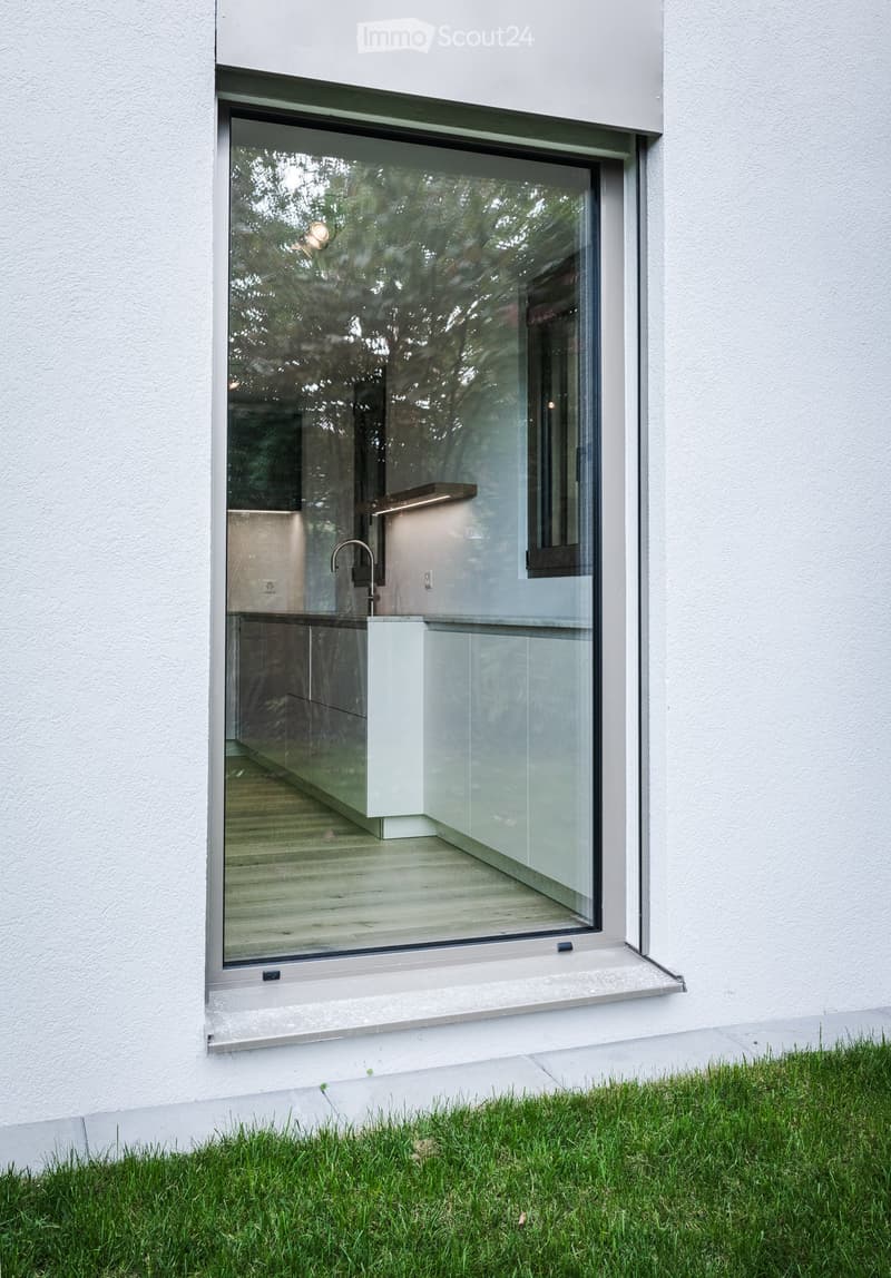 Modern building exterior with large glass window and sleek design, featuring a small outdoor area with wooden flooring and a modern faucet.