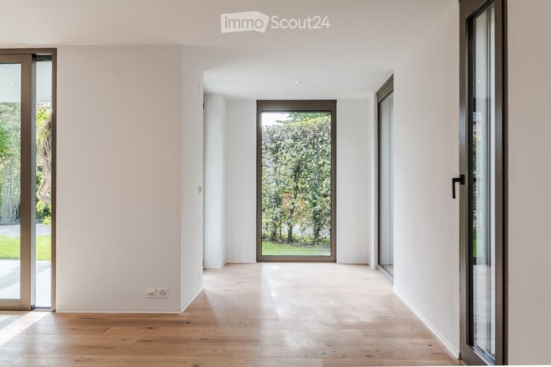 Empty hallway with light wooden flooring, white walls, and large glass doors leading to a garden.