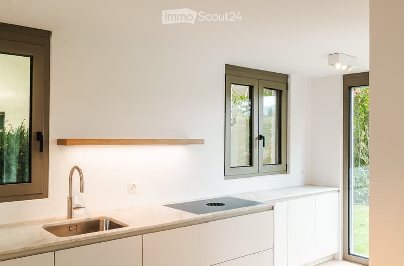 Modern kitchen with white cabinets, marble countertops, a stainless steel sink, and a sleek faucet. Features large windows and a built-in shelf.