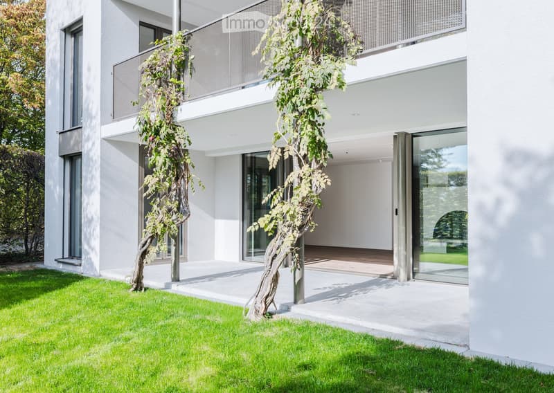 Modern white building with large glass windows and a balcony, surrounded by a well-maintained lawn and a small tree.