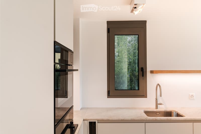 Modern kitchen with white walls, light-colored countertops, a stainless steel sink, a modern faucet, and a built-in oven. A large window with a brown frame offers a view of greenery outside.