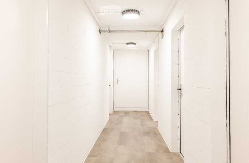 A clean, modern hallway with white brick walls, recessed lighting, and a smooth concrete floor. The hallway features several closed doors and a coat rack with hooks on the left wall.