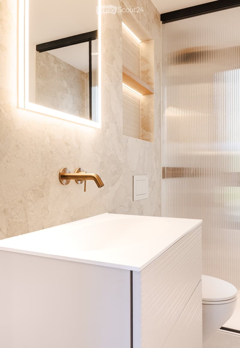 Modern bathroom with a white countertop sink, gold faucet, and a sleek, transparent glass shower enclosure.