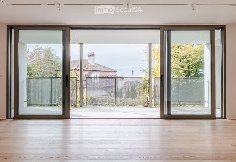 Empty room with light wooden flooring and large sliding glass doors leading to a balcony or outdoor area. The space is well-lit with recessed ceiling lights.