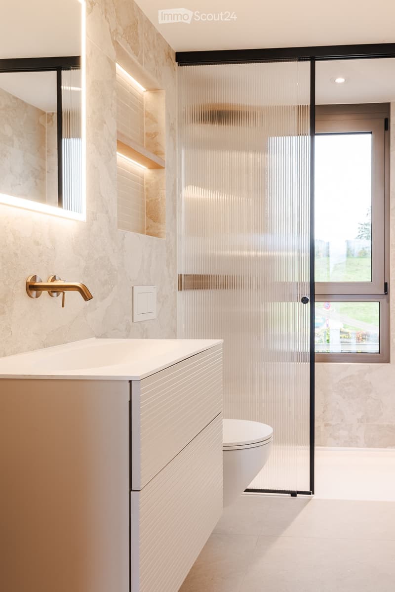 Modern bathroom with a white sink, gold faucet, white toilet, and a sleek glass shower enclosure. The walls are tiled in a marble-like pattern, and there is a window providing natural light.