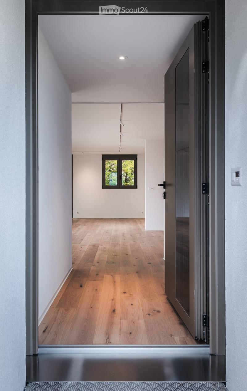 Modern hallway with wooden flooring, white walls, and recessed lighting. Features a large window at the end and a closed door on the right side.