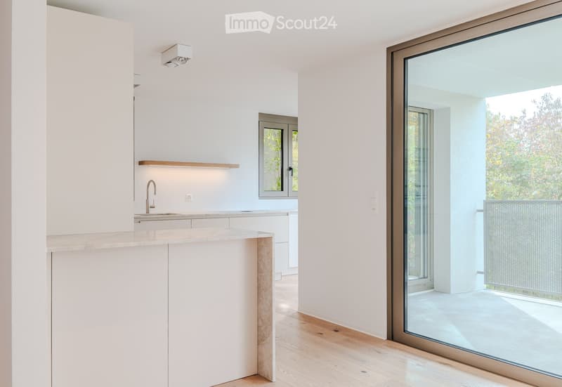 Modern kitchen with white cabinets, a central island, and a large window overlooking a balcony. The space features a sleek, minimalist design with a wooden floor and a marble countertop.