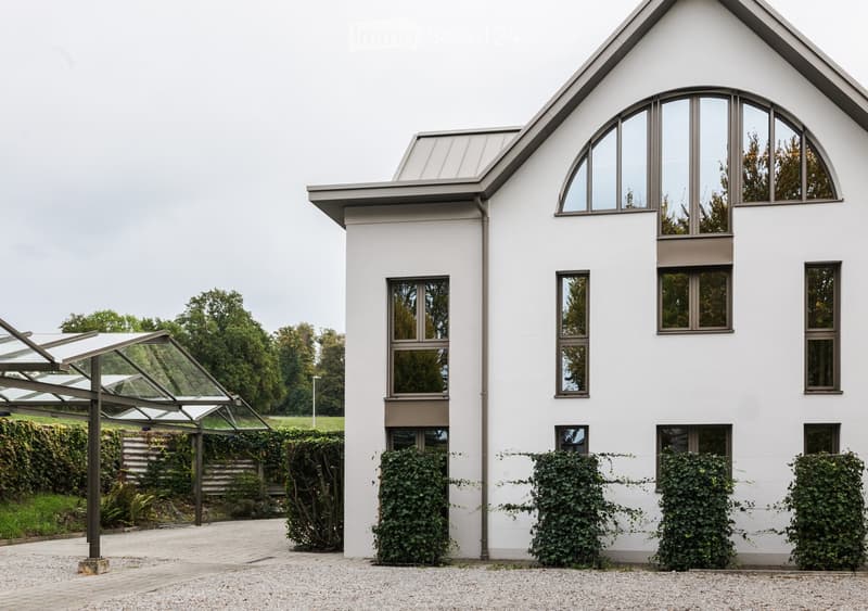 Modern two-story house with white exterior, large windows, and a covered entrance area. The house features a sloped roof and is surrounded by greenery and a paved driveway.