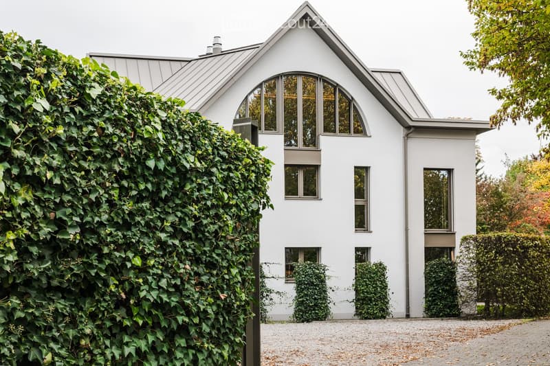 Modern white house with a steeply pitched roof, large arched windows, and a well-maintained hedge on one side.