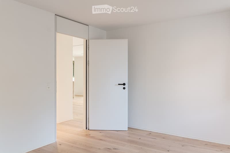 Empty room with light wooden flooring and white walls, featuring a sliding door that opens to another room.