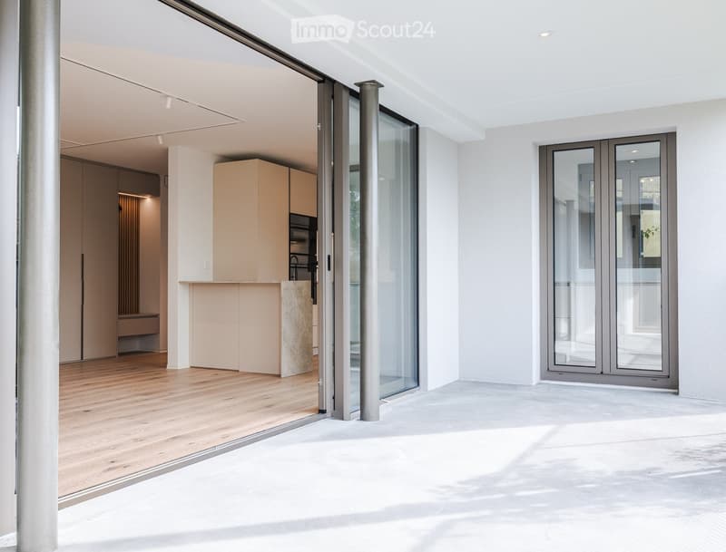 Modern, minimalist kitchen with light wood flooring, cream-colored cabinets, and a stone countertop. Features large glass sliding doors leading to an outdoor space.