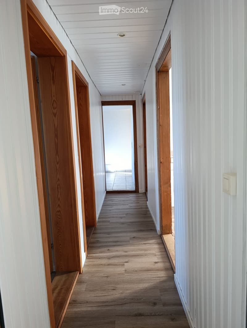 A long, narrow hallway with wooden flooring, white paneled walls, and several wooden doors on both sides. The ceiling is white with recessed lighting, and the hallway leads to an open area at the end.