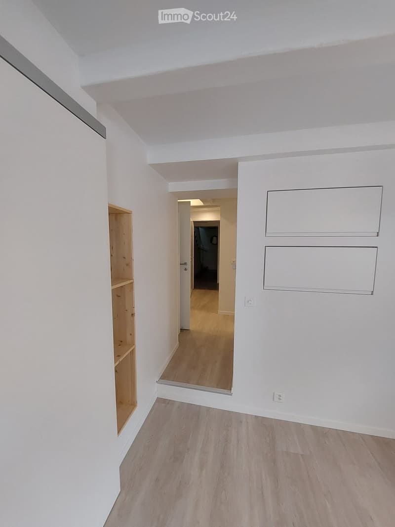 Empty hallway with white walls, light wooden flooring, and a small wooden shelf on the left side. The hallway leads to another room with a visible door and light fixture.