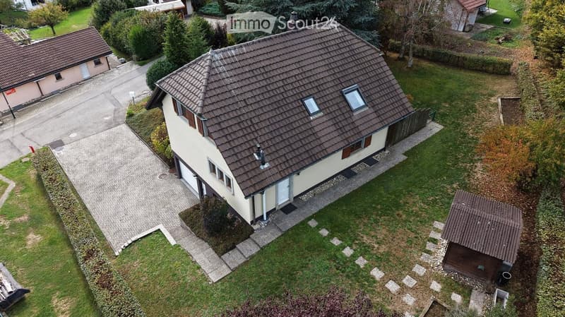 A two-story house with a brown tiled roof, surrounded by a well-maintained garden and driveway. The house features multiple windows and a small wooden shed in the yard.