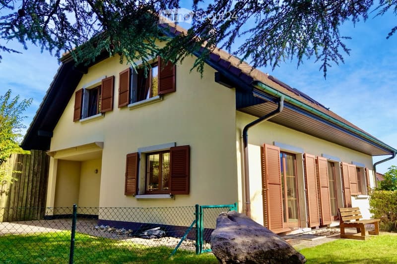 Two-story house with light yellow walls, brown shutters, and a sloped roof. Features include a fenced front yard, a large rock, and a wooden bench near the entrance.