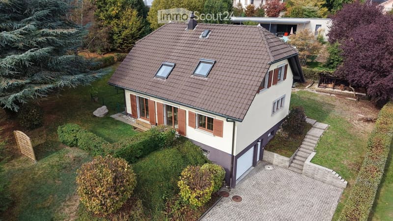 A two-story house with a brown tiled roof, white and beige exterior, multiple windows with shutters, and a well-maintained garden with trees and shrubs.