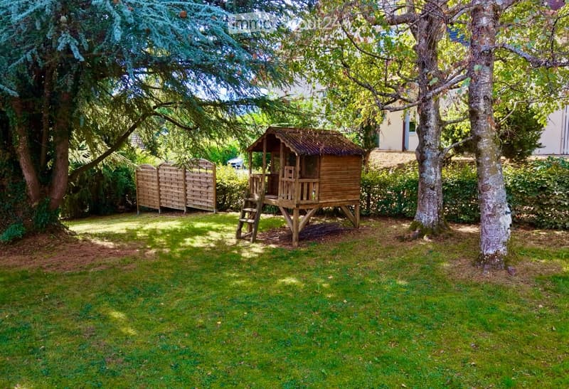 A well-maintained garden with a wooden playhouse, surrounded by trees and a wooden fence, featuring a grassy lawn and a peaceful outdoor setting.