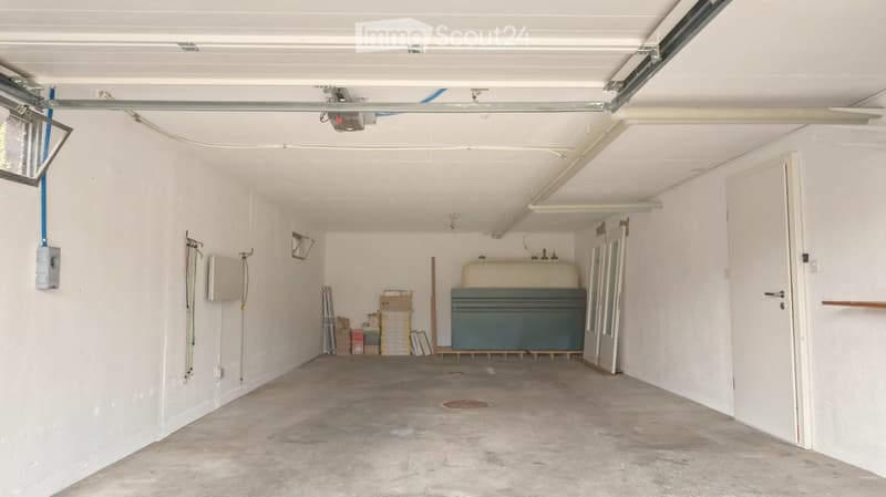 Empty garage with white walls, concrete floor, and storage items in the background.